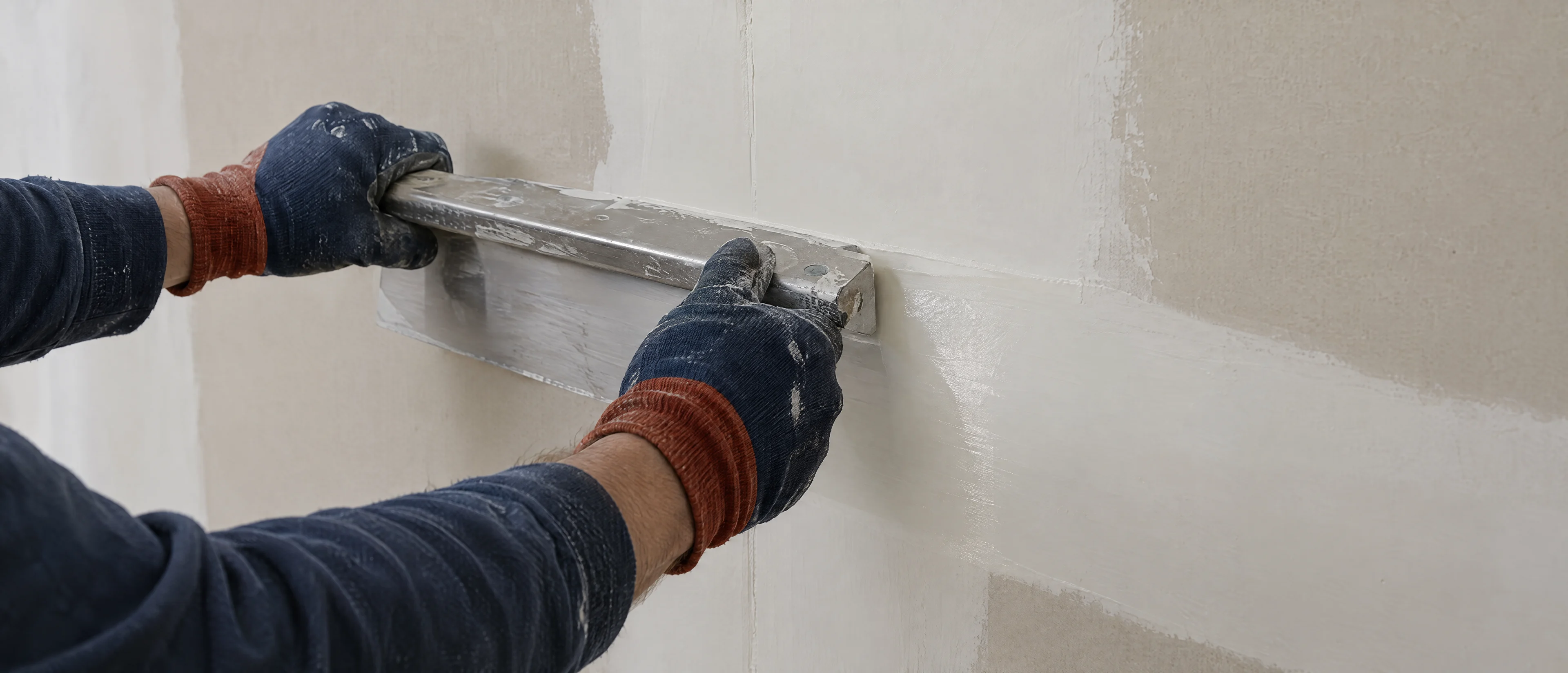 Hände ziehen Feinspachtel mit einer breiten Glättkelle über eine Gipskartonwand